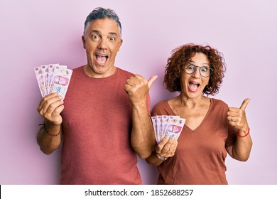 Beautiful Middle Age Couple Holding 50 Mexican Pesos Banknotes Pointing Thumb Up To The Side Smiling Happy With Open Mouth 