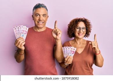 Beautiful Middle Age Couple Holding 50 Mexican Pesos Banknotes Smiling With An Idea Or Question Pointing Finger With Happy Face, Number One 