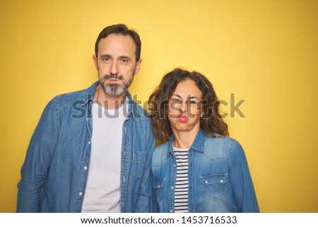 Beautiful middle age couple together wearing denim shirt over isolated yellow background puffing cheeks with funny face. Mouth inflated with air, crazy expression.