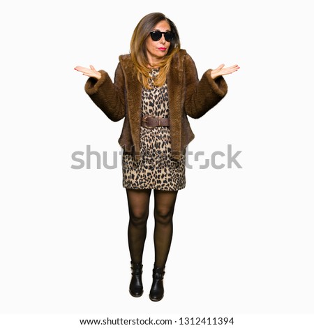 Beautiful middle age elegant woman wearing sunglasses and mink coat clueless and confused expression with arms and hands raised. Doubt concept.