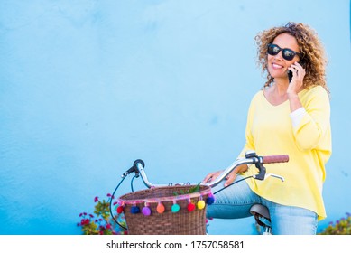 Beautiful Middle Age Healthy Young Woman Do A Phone Call Sit Down On A Vintage Coloured Happy Bike And Blue Background Wall - Colors And Happy People Lifestyle Concept