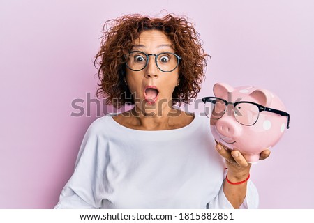 Beautiful middle age mature woman holding piggy bank with glasses scared and amazed with open mouth for surprise, disbelief face 