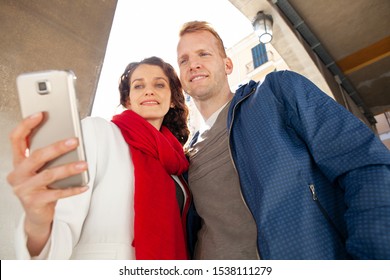 Beautiful Middle Age Tourist Couple On Sunny Winter City Break Holiday Sightseeing Together, Using Technology Smartphone Smiling Outdoors. Fun Travel Vacation, People Leisure Recreation Lifestyle.