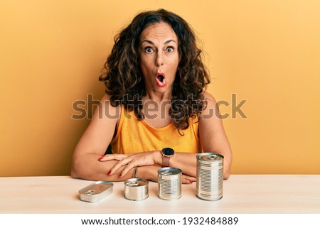Beautiful middle age woman with canned food scared and amazed with open mouth for surprise, disbelief face 