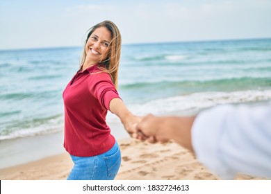 Beautiful Middle Age Woman Holding Hands With Husband At The Beach, Leading The Way Happy Smiling In Love