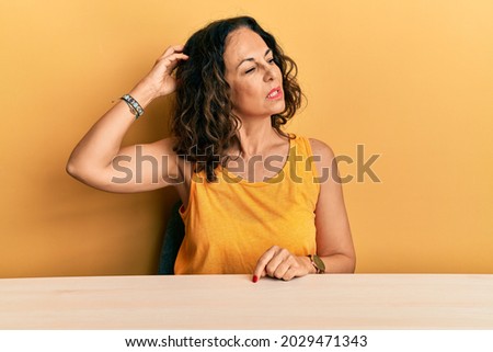 Beautiful middle age woman wearing casual clothes sitting on the table confuse and wondering about question. uncertain with doubt, thinking with hand on head. pensive concept. 