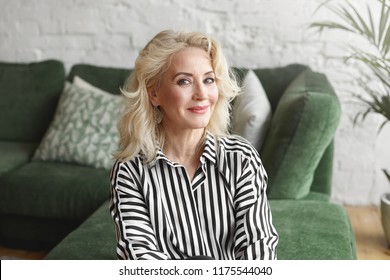 Beautiful Middle Aged 50 Year Old Female Wearing Stylish Elegant Striped Blouse Relaxing At Home, Sitting On Comfortable Green Sofa In Modern Living Room Interior, Looking At Camera With Joyful Smile