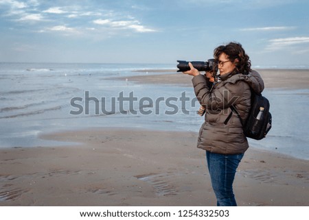 Beautiful, middle aged Brazilian woman with glasses on the beach, holding a camera. She looks through the camera and takes pictures along the ocean. Cold, blue, beautiful day in the Netherlands.