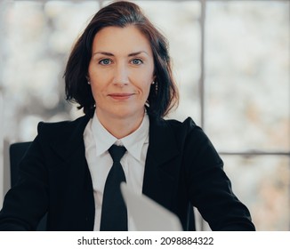 Beautiful Middle Aged Businesswoman Wears Black Suit And Tie Looking At Camera Smiling. Portrait Of Female Director CEO Team Leader Sitting Blurred Background. Coaching And Mentoring Business Concept.