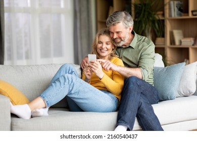 Beautiful Middle Aged Couple Using Mobile Phone And Embracing Together At Home, Happy Mature Spouses Relaxing On Couch With Modern Smartphone, Browsing New App Or Shopping Online, Copy Space - Powered by Shutterstock