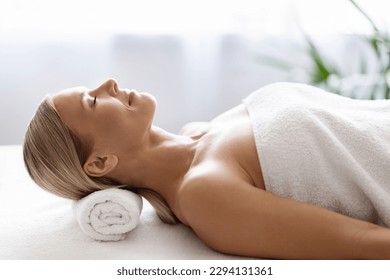 Beautiful Middle Aged Female Resting Before Beauty Treatment In Spa Salon, Side View Shot Of Attractive Mature Woman Lying On Table With Eyes Closed, Using Rolled Towel As Pillow, Copy Space - Powered by Shutterstock