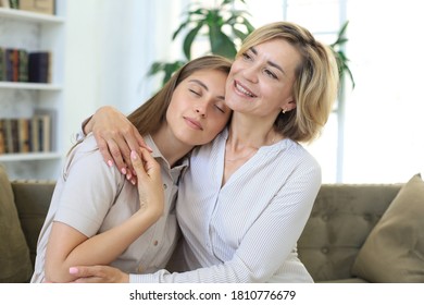 Schöne Mutter mittleren Alters und ihre erwachsene Tochter umarmt und lächelt, während sie zu Hause auf dem Sofa sitzt.