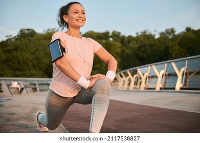 Beautiful middle aged sportswoman with earphones and smartphone holder enjoying morning workout at sunrise, stretching legs, doing lunges, squatting before jogging. Female athlete working out outdoor - Powered by Shutterstock