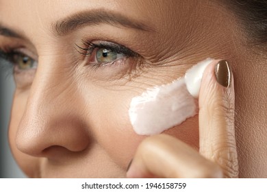 Beautiful middle aged woman applying anti-aging cream on her face on gray 