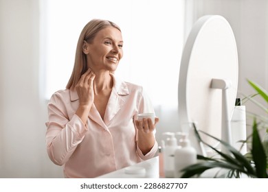 Beautiful middle aged woman applying moisturizing cream on her neck while sitting near mirror at home, attractive mature female enjoying domestic beauty routine, holding jar with nourishing lotion