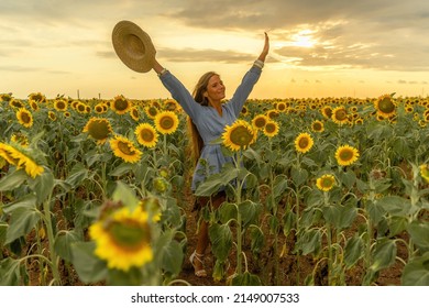 Beautiful Middle Aged Woman Looks Good In A Hat Enjoying Nature In A Field Of Sunflowers At Sunset. Summer. Attractive Brunette With Long Healthy Hair.