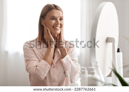 Beautiful Middle Aged Woman Touching Face And Looking In Mirror At Home, Happy Mature Attractive Lady Enjoying Her Flawless Skin, Smiling Female Making Anti-Aging Beauty Routine, Closeup