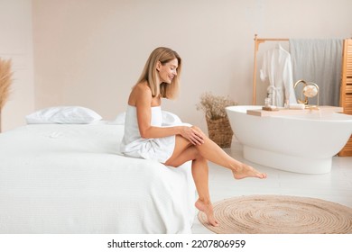 Beautiful middle aged woman in towel touching her leg with soft skin after depilation, sitting on bed at home, copy space. Mature lady after hair removal procedure, full length