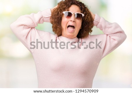 Beautiful middle ager senior woman wearing pink sweater and sunglasses over isolated background Crazy and scared with hands on head, afraid and surprised of shock with open mouth