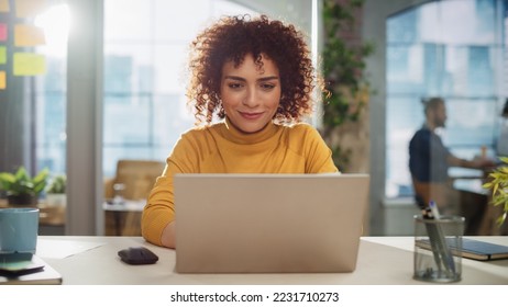 Beautiful Middle Eastern Manager Sitting at a Desk in Creative Office. Young Stylish Female with Curly Hair Using Laptop Computer in Marketing Agency. Colleagues Working in the Background