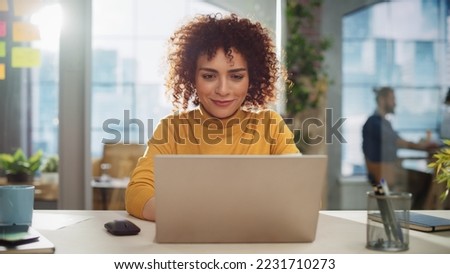 Beautiful Middle Eastern Manager Sitting at a Desk in Creative Office. Young Stylish Female with Curly Hair Using Laptop Computer in Marketing Agency. Colleagues Working in the Background