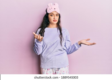 Beautiful middle eastern woman wearing sleep mask and pajama clueless and confused expression with arms and hands raised. doubt concept. 