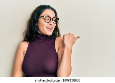 Beautiful middle eastern woman wearing casual clothes and glasses pointing thumb up to the side smiling happy with open mouth 
