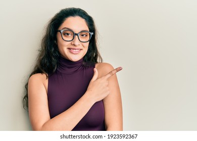 Beautiful middle eastern woman wearing casual clothes and glasses smiling cheerful pointing with hand and finger up to the side 