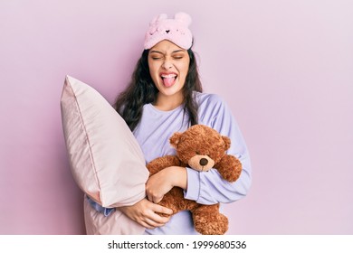 Beautiful middle eastern woman wearing sleep mask hugging pillow sticking tongue out happy with funny expression.  - Powered by Shutterstock