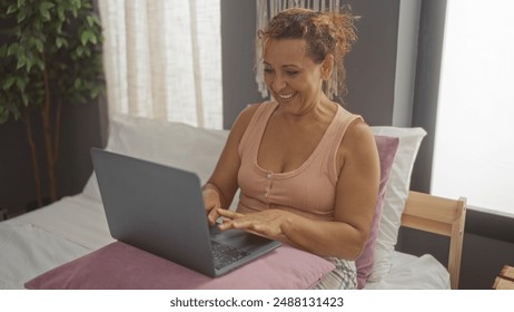 A beautiful middle-aged hispanic woman uses a laptop while sitting on her bed in a cozy bedroom, radiating happiness. - Powered by Shutterstock