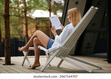 beautiful middle-aged lady with blonde hair sitting on an armchair on the terrace of an A-frame house on a sunny summer afternoon with a smile on her face reading an interesting book - Powered by Shutterstock