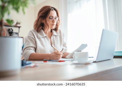 Beautiful middle-aged woman in glasses counting family business budget gazing at monthly bills and documents at home living room office. Small business home finances, money savings concept image - Powered by Shutterstock