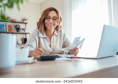 Beautiful middle-aged woman in glasses and paper bills joyfully laughing while calculating revenue grows at home office. Small business, successful investment, bull market, money savings concept image - Powered by Shutterstock