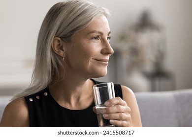 Beautiful middle-aged woman hold glass of clear water smile look into distance, enjoy healthy morning routine, drink still and mineral water start new day, close up. Lifestyle, beauty skin, healthcare