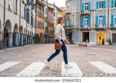 Beautiful middle-aged woman with long blonde hair wearing jeans, beige sweater, sunglasses and leather handbag crossing street on pedestrian crossing in European city on sunny autumn day. Side view - Powered by Shutterstock