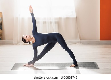 A beautiful middle-aged woman practicing yoga at home, doing extended triangle exercises, a trainer in dark blue sportswear stands in the Utthita Trikonasana pose on the mat.