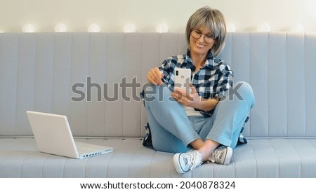 A beautiful middle-aged woman is sitting on the couch and having fun chatting and laughing with someone on the phone. The concept of online communication on a laptop and phone remotely.