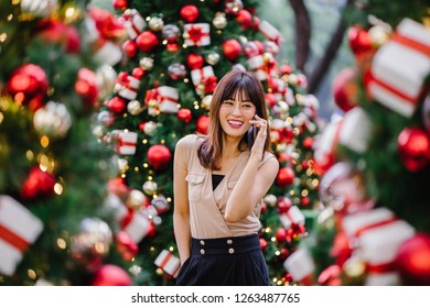A beautiful middle-aged woman is talking happily on her smartphone and smiling as she stands amidst beautifully decorated Christmas trees during the day. 