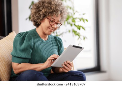 Beautiful middle-aged woman using a computer tablet to read a book and surf internet. Modern technologies used by elderly people. Mature woman using digital tablet at home - Powered by Shutterstock