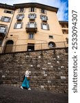 Beautiful middle-aged woman wearing white t-shirt, long turquoise skirt, handbag and sunglasses walking down street against background of stone wall in old town in summer. Side view. Bergamo, Italy.