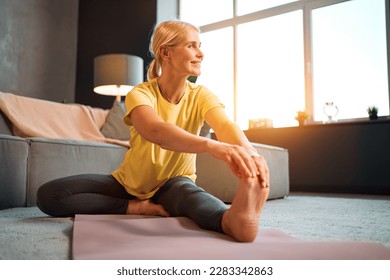 A beautiful middle-aged woman in a yellow t-shirt and gray leggings is doing exercises while sitting on a pink rug and looking away in a room at home. Sports and health. - Powered by Shutterstock