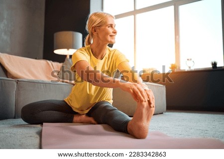 A beautiful middle-aged woman in a yellow t-shirt and gray leggings is doing exercises while sitting on a pink rug and looking away in a room at home. Sports and health.