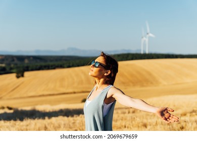 Beautiful Middle-aged Young Woman Wearing Sunglasses Breathing Air In Cereal Field With Wind Farm In Background. Healthy Life Style