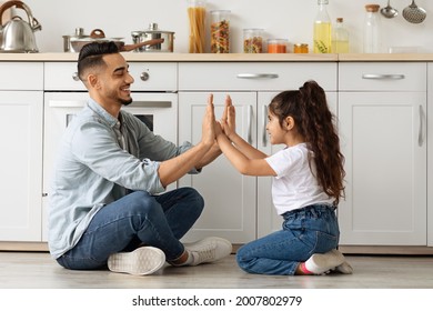 Beautiful middle-eastern family father and daughter playing at kitchen, sitting on floor and clapping hands. Cheerful arab dad and little female kid having fun together at home, side view, copy space - Powered by Shutterstock