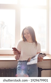 Beautiful Millenial Woman Using Mobile Phone And Smiling While Standing Near Window. Natural Morning Light. Chatting Or Shopping Online