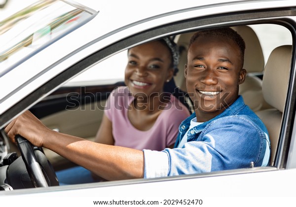 Beautiful millennial afro american couple posing in\
their new auto, cheerfully smiling at camera while making test\
drive, side view, closeup. Happy black man and woman going summer\
vacation by car