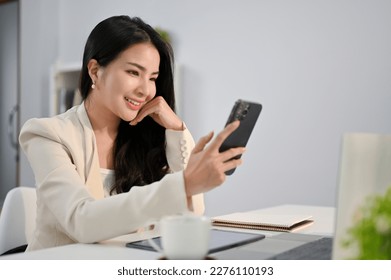 Beautiful millennial Asian businesswoman hand on chin, looking at her phone screen, using her phone, scrolling on social media or using mobile app at her desk.