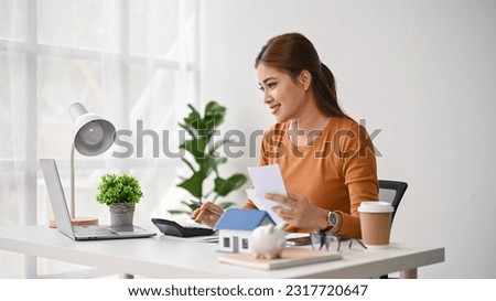 A beautiful millennial Asian woman sits at her desk, paying her bills online on her laptop. electricity bills, monthly expenses, savings