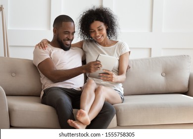 Beautiful millennial biracial wife sit on husband lap knees watching video on tablet together, smiling african American young couple relax on comfortable sofa using pad, enjoy weekend at home - Powered by Shutterstock