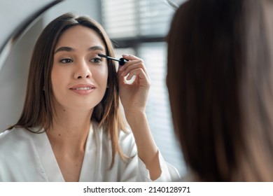 Beautiful Millennial Lady Doing Daily Makeup Near Mirror In Bathroom, Attractive Young Woman Applying Mascara On Eyelashes, Holding Brush And Smiling To Her Reflection, Selective Focus, Closeup